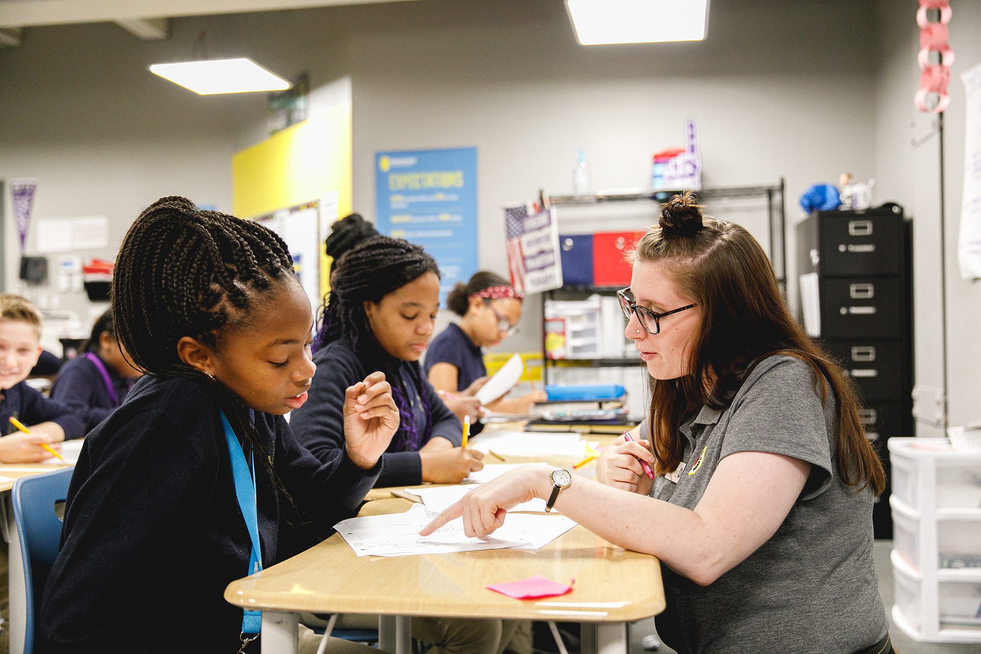 americorps volunteer in a classroom full of students helping a girl at her desk with a problem