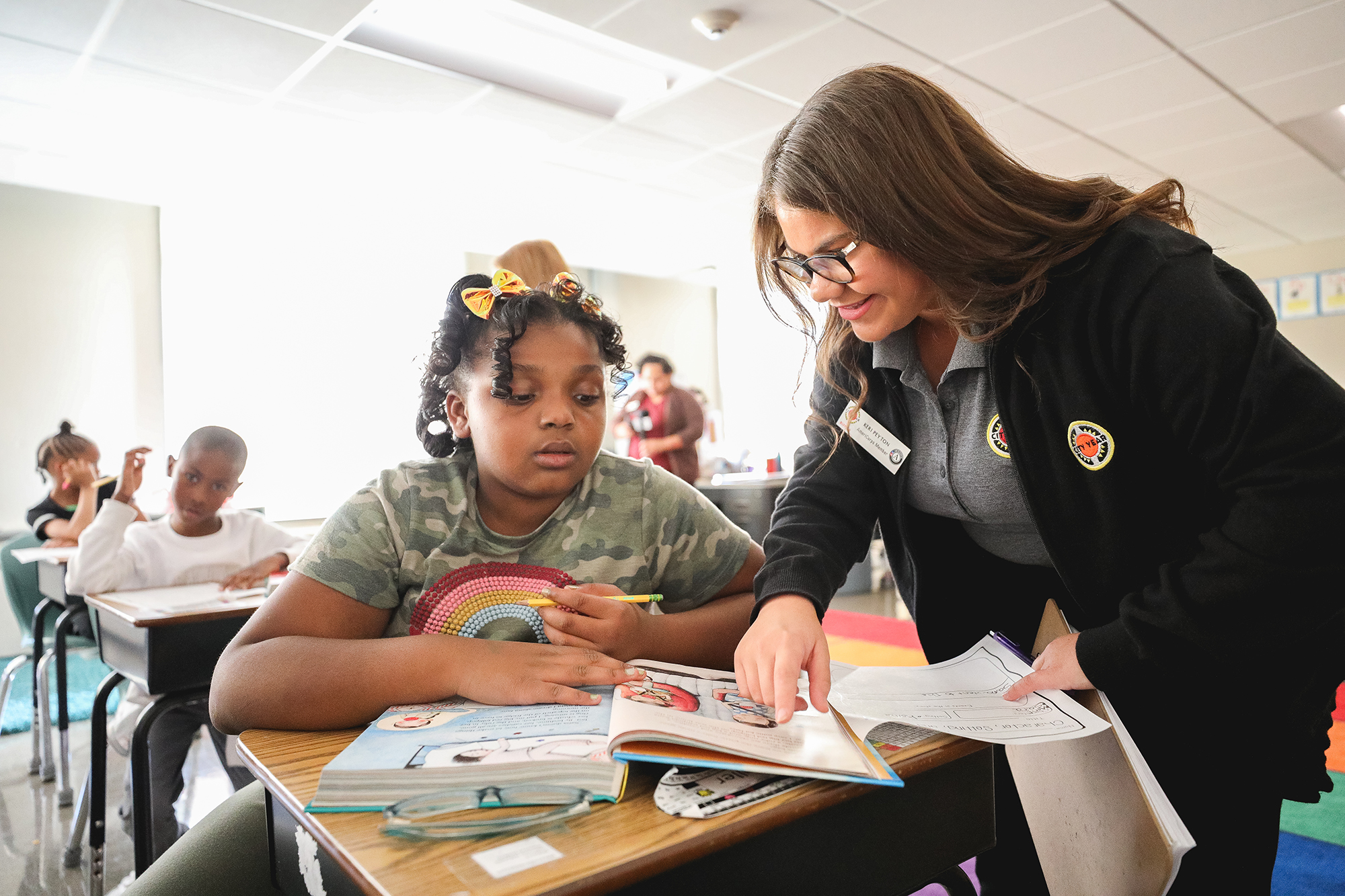 americorps volunteer helping younger girl at her desk with a book in front of them